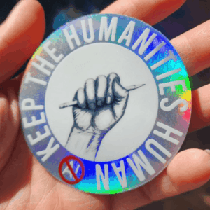 A hand holds a round sticker with a holographic border. The sticker shows a raised fist gripping a pencil and the words “KEEP THE HUMANITIES HUMAN”; a red circle with a slash symbol is in the bottom left.