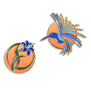 Two enamel pins are shown. One pin depicts a blue hummingbird with outstretched wings in front of an orange circle. The other pin shows a blue flower on a green stem set against an orange circle.