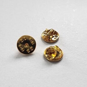 Three small gold-colored metal pin backs are shown on a white surface. Two are positioned with the clasp side visible, and one displays the bottom with six spikes around a central hole.