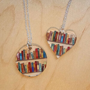 Two silver necklaces with pendants featuring colorful books on shelves; one pendant is round and the other is heart-shaped, both resting on a light wood surface.