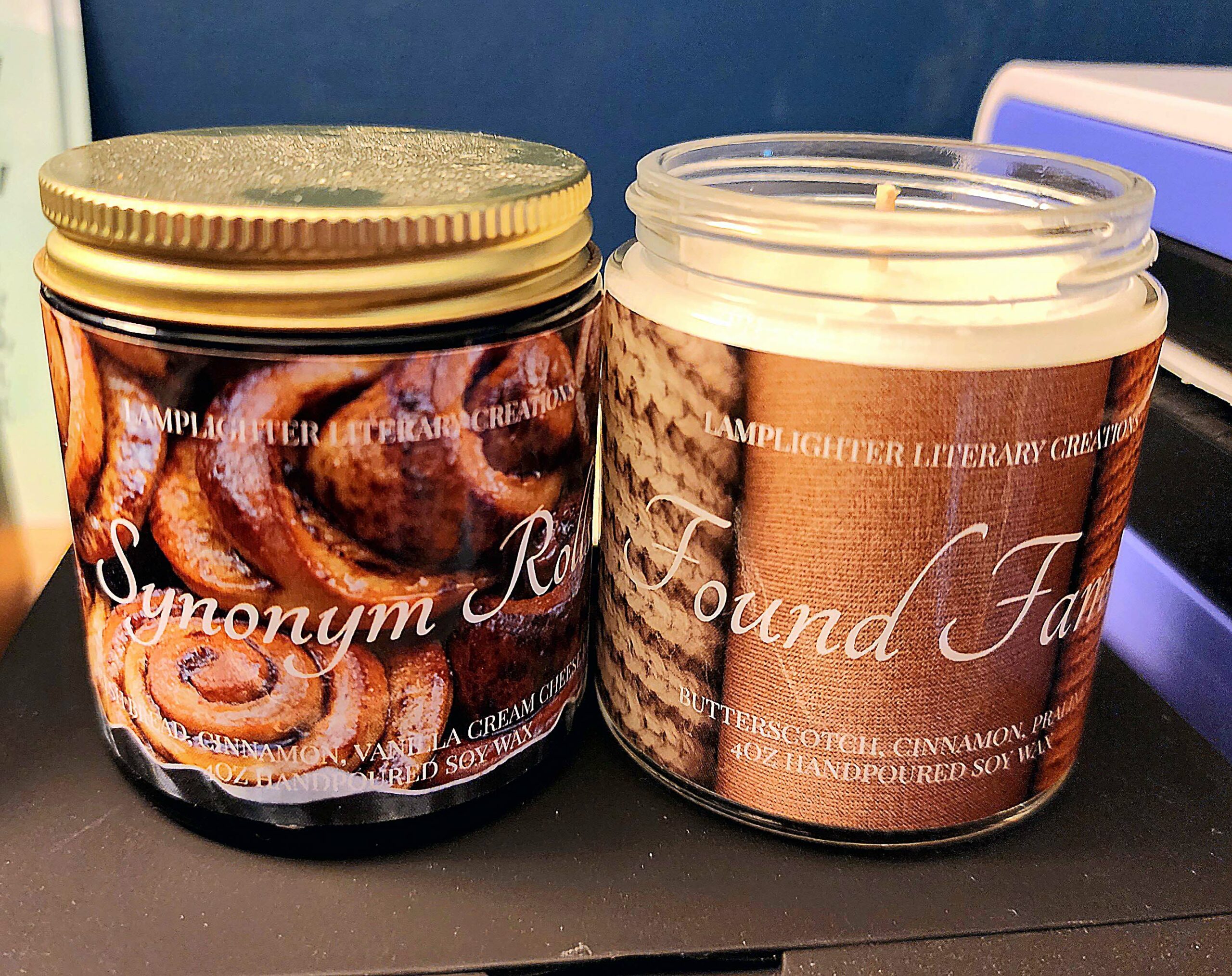 Candles for Booklovers: Two glass jars with printed labels sit side by side—one labeled "Synonym Roll" with a cinnamon roll design, the other "Found Family" featuring a cozy, textured pattern.